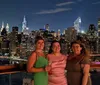 Three smiling women pose together at night with a vibrant city skyline illuminated in the background