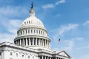 The image shows a close-up view of the United States Capitol dome with the American flag flying nearby against a partly cloudy sky.